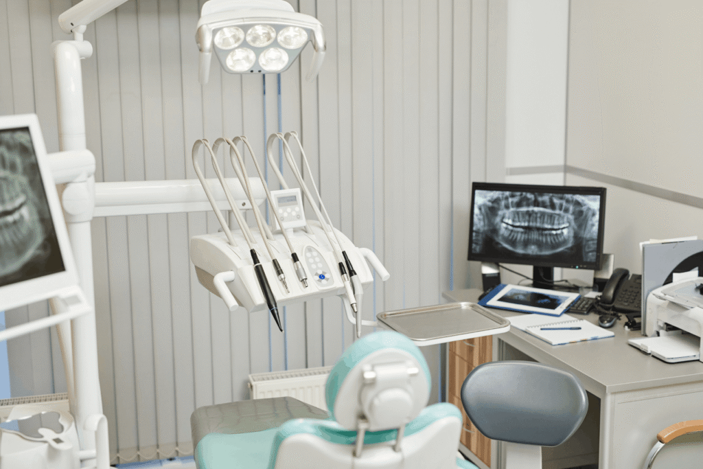 Dental office with a chair, overhead light, monitor showing X-rays, and various dental tools. The room is bright and organized, conveying a clinical atmosphere.