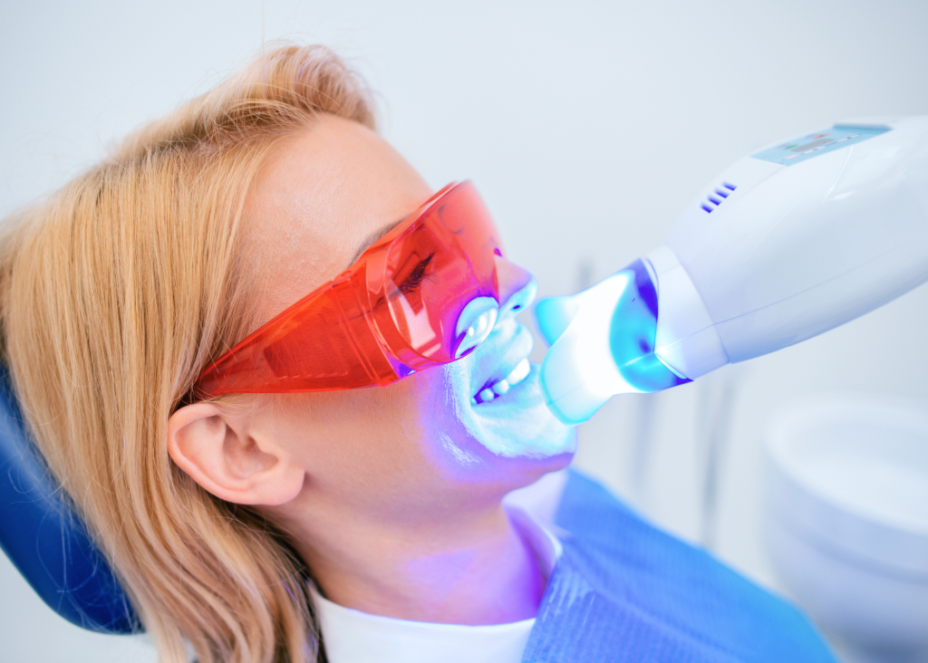 A woman in a dental chair receives teeth whitening treatment, wearing red protective glasses. A blue light device illuminates her mouth.