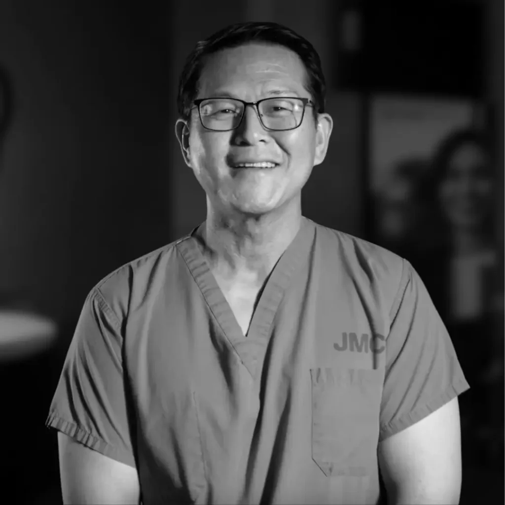 A man wearing glasses and medical scrubs with JMC on the chest smiles at the camera. The image is in black and white, and the background is softly blurred.