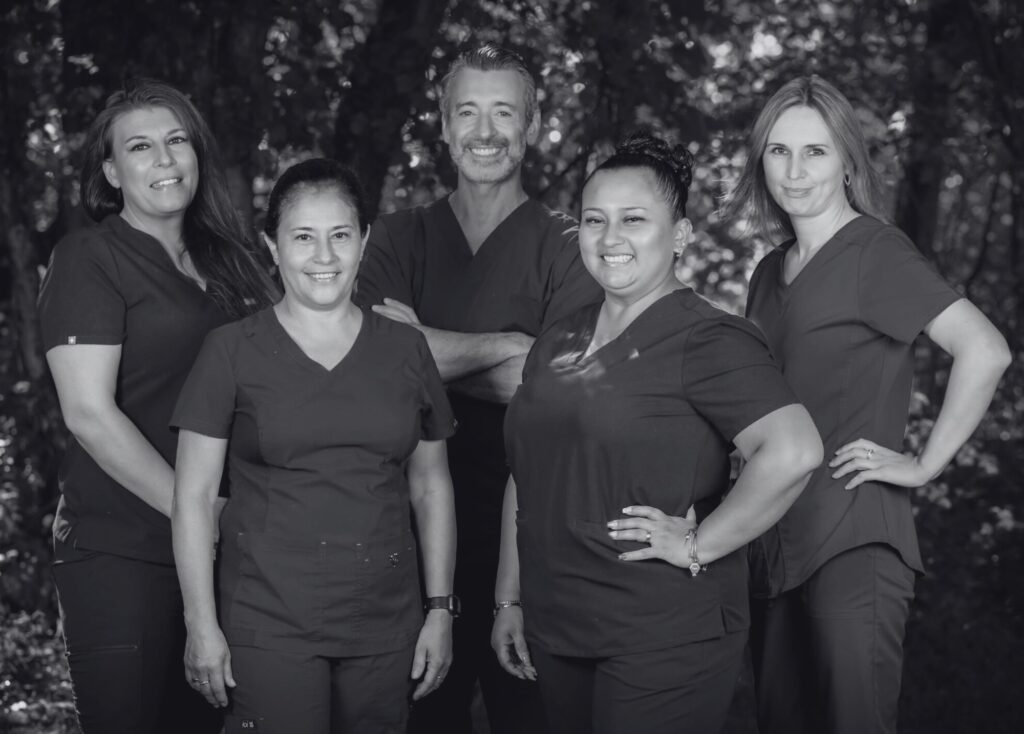 A group of five people in medical scrubs stands outdoors, smiling confidently. The black and white image conveys a professional yet approachable tone.