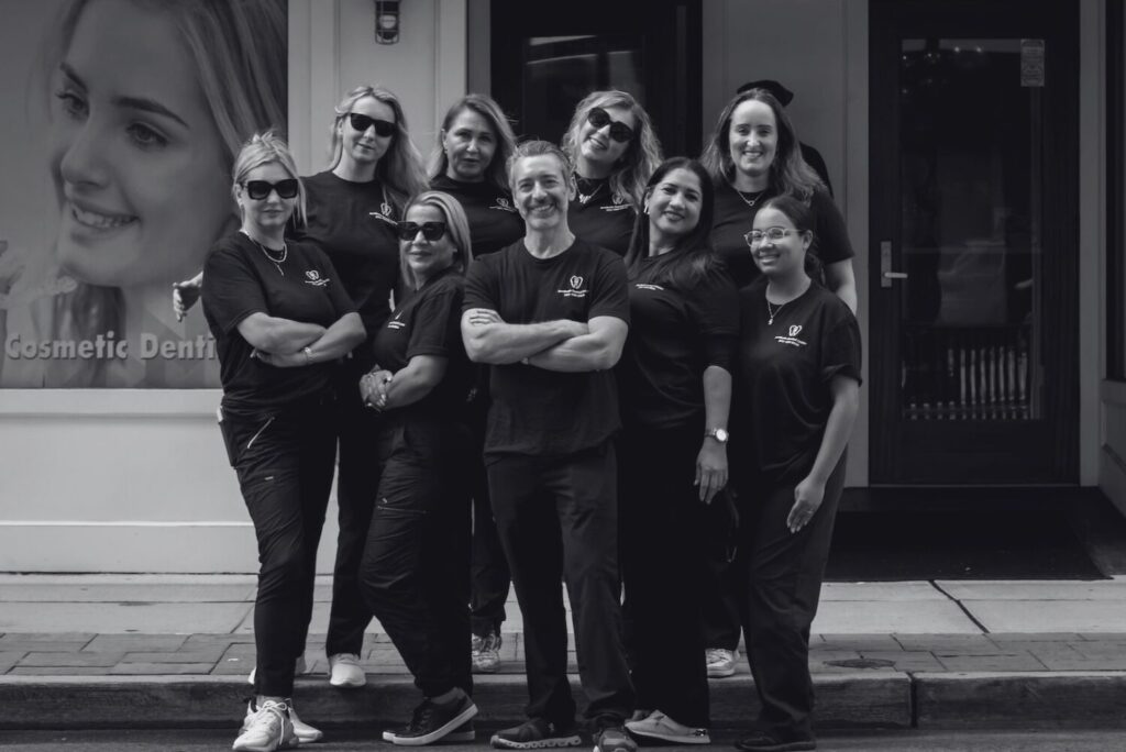 A group of nine people in matching dark shirts pose together and smile outside a building, with a large poster of a womans face in the background. The photo is black and white.